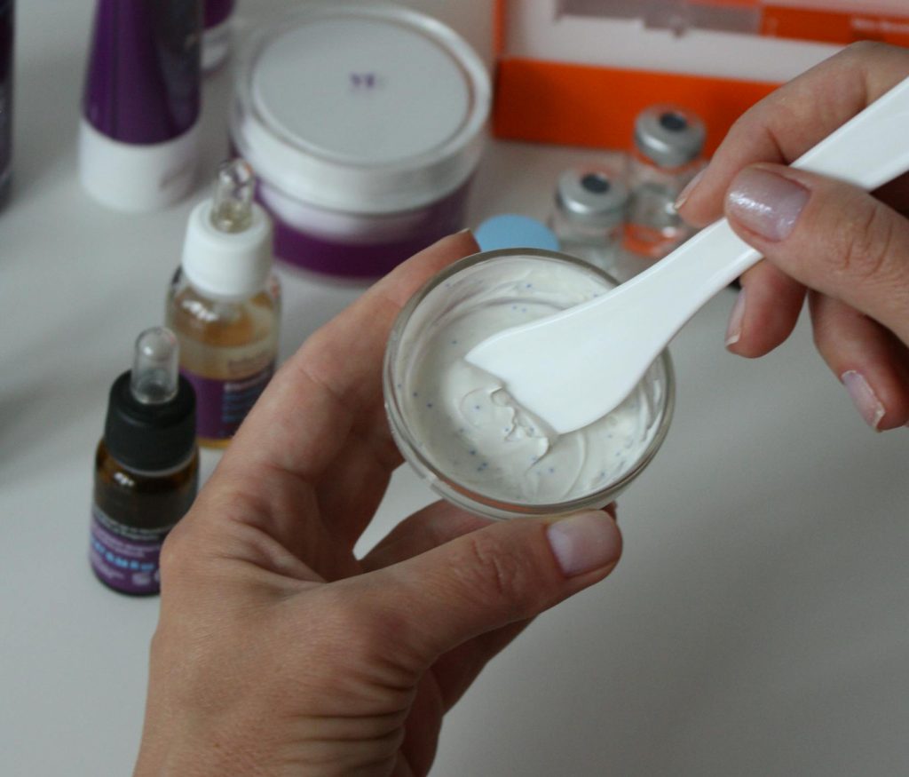 Close-up of hands mixing beauty product in glass jar, surrounded by cosmetics on white background.